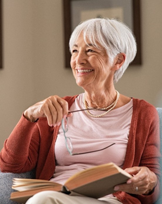 Woman with white hair holding glasses reading in a gray chair smiling